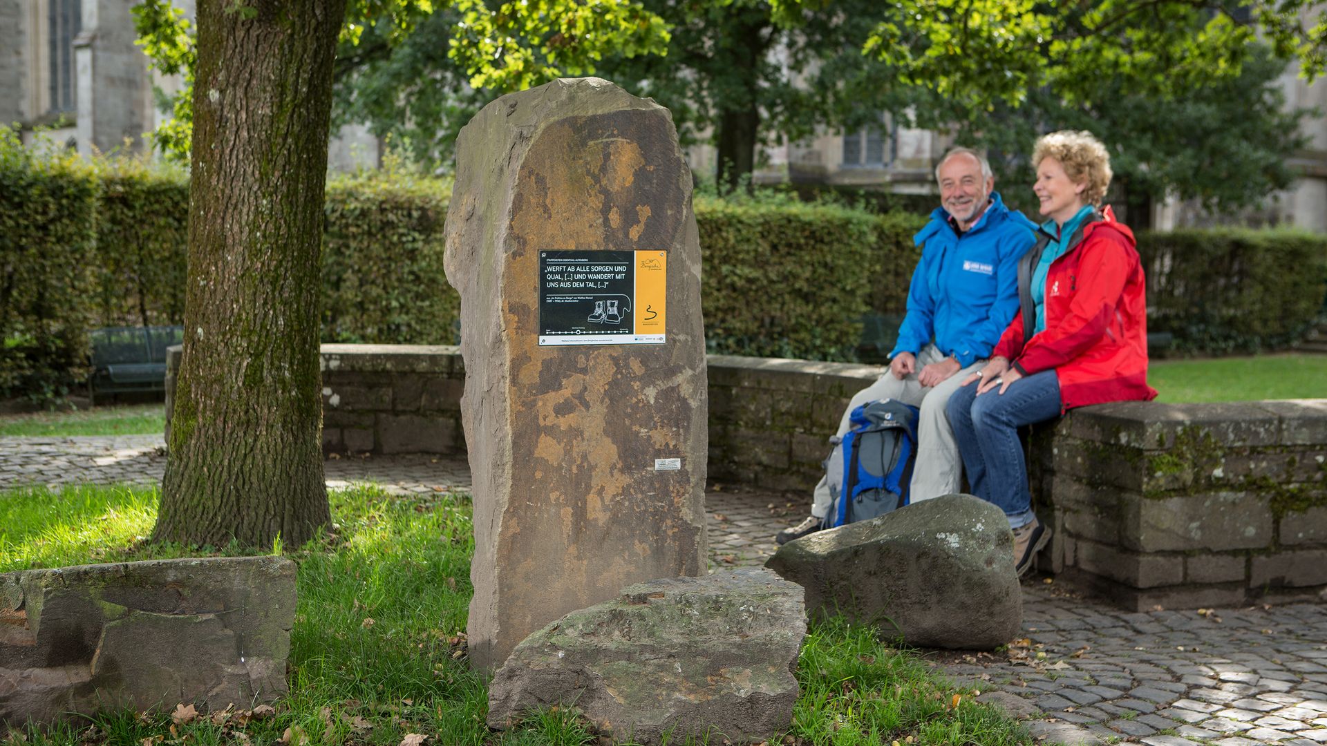 Ein Mann und eine Frau in Wanderkleidung sitzen auf einer kleinen Mauer. Im Vordergrund ist ein Grauwackestein mit einem Schild zu sehen, im Hintergrund Bäume und ein Teil der Außenwamd des Altenberger Doms. 