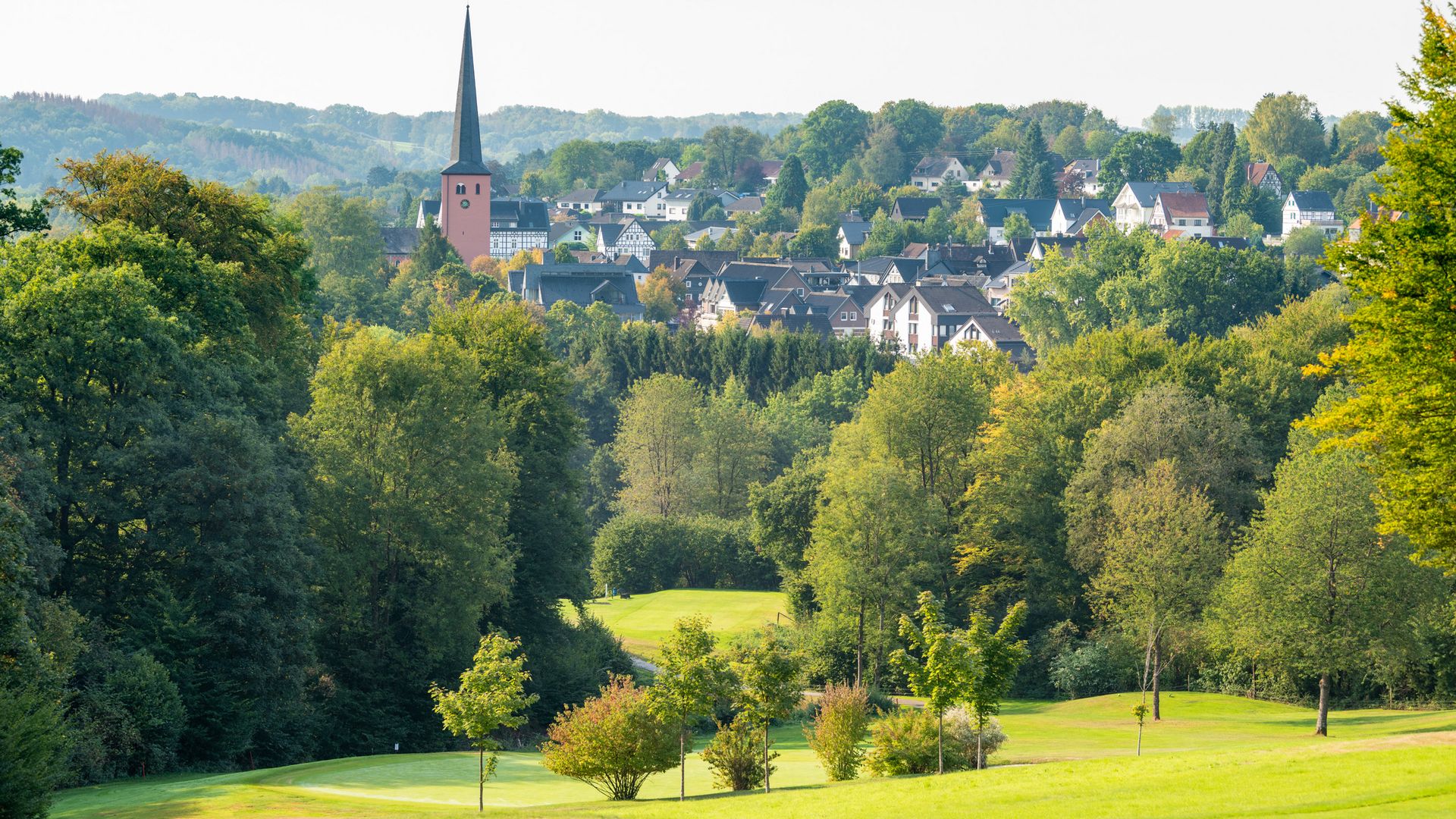 Blick vom Golfplatz auf das Ortszentrum von Much mit einer rosafarbenen Kirche und vielen Wohnhäusern. Der Ort ist vob zahlreichen Bäumen umgeben. 