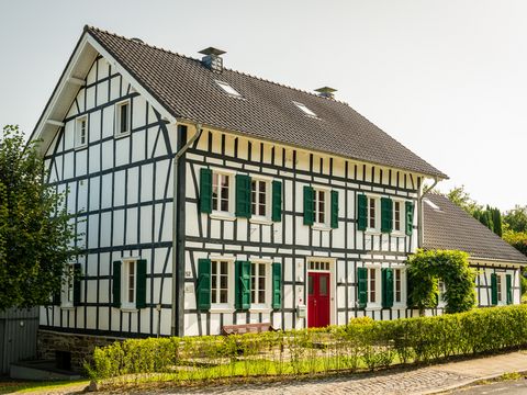Typisch bergisches Fachwerkhaus, mit schwarzen Balken, weißem Gefache, grünen Schlagläden und einer roten Hausür, davor eine kleine hellgrüne Hecke