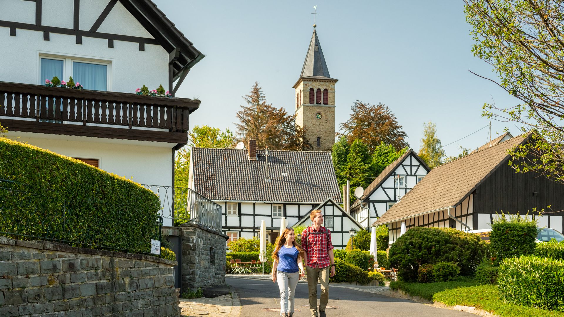 Ein junges Paar wandert auf einer Straße, im Hintergrund stehen Fachwerkhäuser, hinter denen ein Kirchturm emporragt.