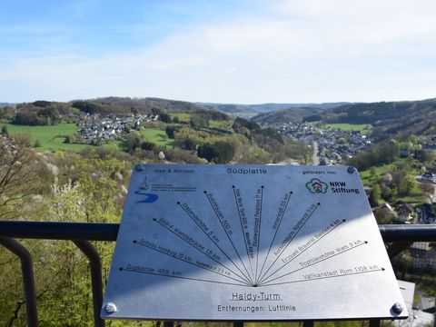 Am Geländer der Aussichtsplattform vom Haldyturm ist eine Tafel angebracht, die Richtungen und Entfernungen zu anderen Orten anzeigt. Im Hindergrund schaut man über das Aggertal.