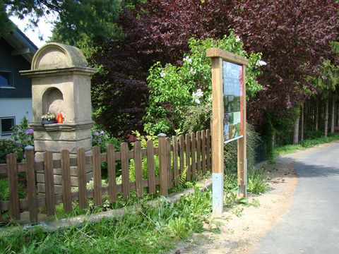An einer Straße steht vor einem Zaun eine Infotafel des Overather Pilgerweges. Hinter dem Zaun steht eine Gedenkstätte mit roten Kerzen.