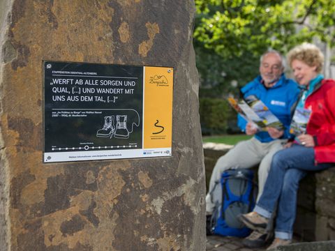 Im Vordergrund ist ein Grauwackestein mit einem Schild zu sehen, im Hintergrund ist unscharf ein Mann und eine Frau in wanderkleidung sichtbar die auf einer Mauer sitzen und in eine Wanderkarte schauen. 