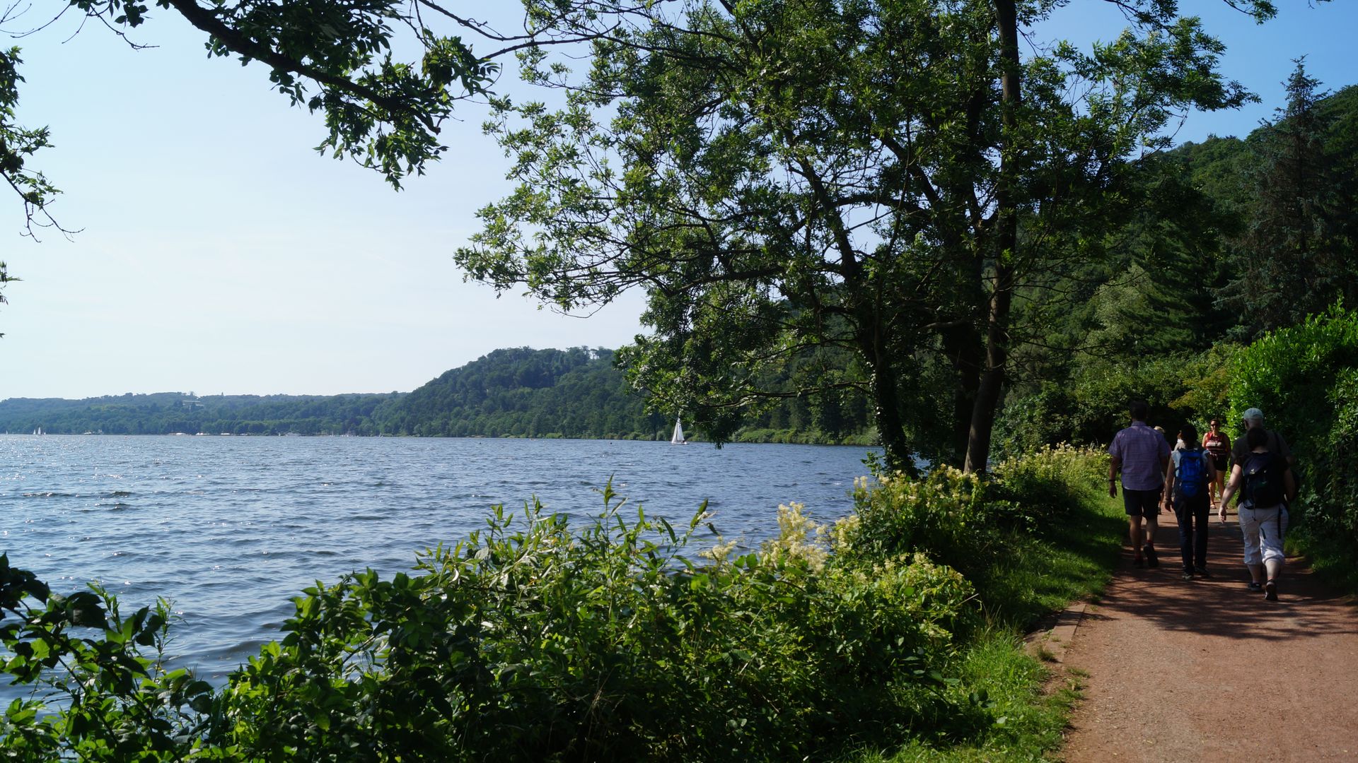 Wanderweg am Ufer des Baldeneysees mit einer Wandergruppe auf der rechten Seiten und dem Blick auf das Ufer und den See bei schönem Wetter auf der linken Seite. 