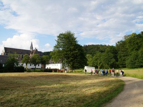 Eine kleine Wandergruppe wandert auf einem Wiesenweg auf ein paar Gebäude vor dem Altenberger Dom zu. 