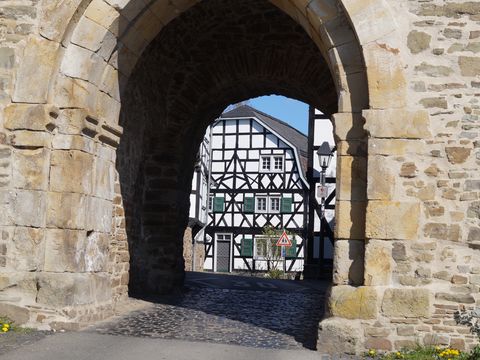 Blick auf ein Fachwerkhaus mit grünen Fensterläden durch den gemauerten Torbogen eines Turmes. 