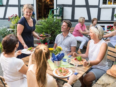 Eine junge Kellnerein serviert vier Personen an einem Holztisch im Biergarten vor einem Fachwerkhaus gerade ihre Gerichte. 