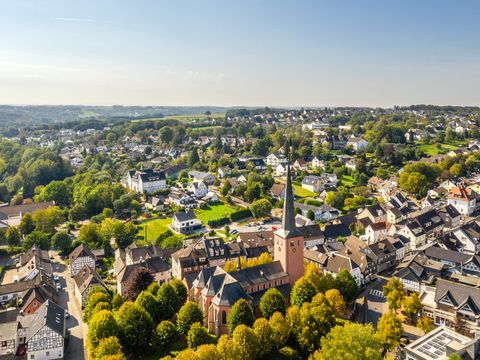 Luftaufnahme des Ortszentrums von Much. Im Zentrum steht eine rosafarbene Kirche, drumherum stehen Wohlhäuser und zahlreiche Bäume. 