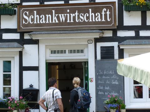 Vor der geöffneten Tür eines Fachwerkhauses mit einem großen Schild "Schankwirtschaft" stehen ein Mann mit einer Kamera übe der Schulder und eine blonde Frau mit einem Rucksack auf dem Rücken.