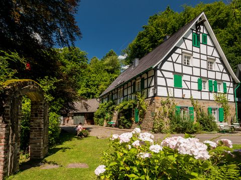 Fachwerkhaus mit grünen Fensterläden in einem blühenden Garten mit einem Torbogen aus Stein am linken Bildrand.