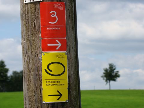 An einem Zaunpfahl sind die Wegemarkierungen des Heimatweges in rot und die des Bergischen Panoramasteiges in gelb angebracht. Im Hintergrund eine grüne Wiese mit einem einzelnen Baum und blauer Himmel mit weißen Wolken.