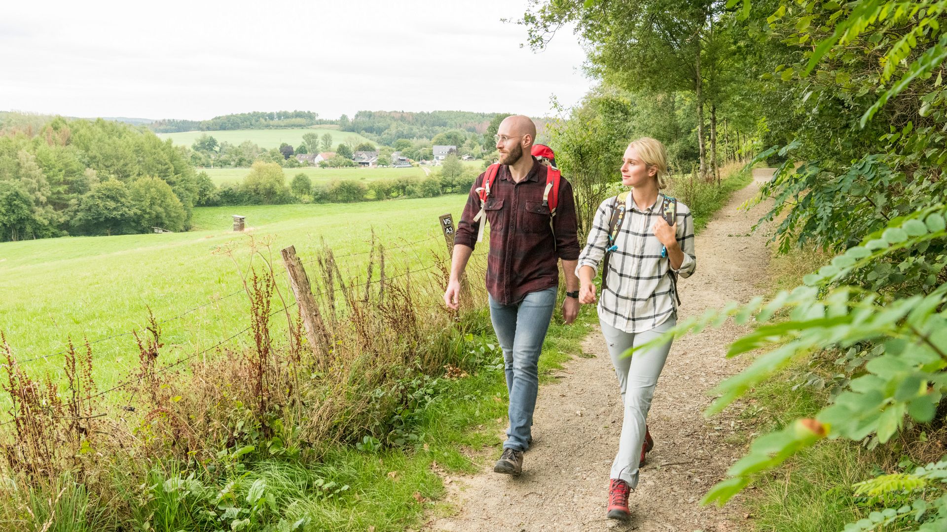 Ein junges Paar wandern über einen Waldweg. Am rechten Wegesrand stehen Bäume und Sträucher, am linken ist eine Wiese mit Fernblick über grüne Hügel und Wälder.