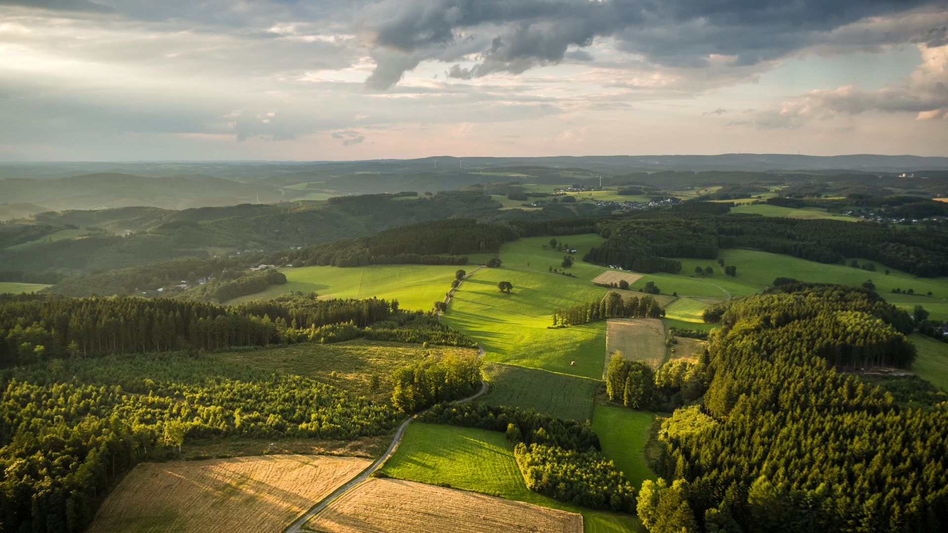 Luftaufnahme der hügeli.gen bergischen Landschaft. Am Himmel sind dunke Wolken zu sehen durch die die Sonne scheint. 