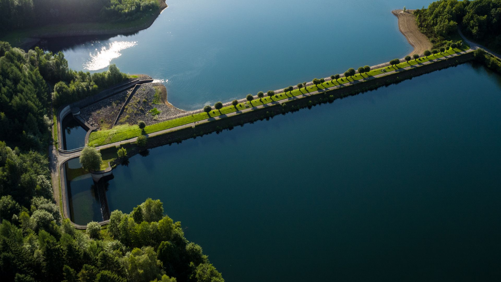 Luftaufnahme eines Staudamms der die Vorsperre von der Talsperre trennt. Au dem Staudamm wachsen Bäume und an beiden Seiten des Damms ist etwas vom Ufer sichtbar. 