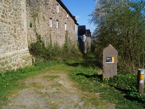 Grauwackestein mit einem Hinweisschild auf den Bergischen Weg steht rechts von einer historischen Mauer auf einer Grünfläche mit Sträuchern