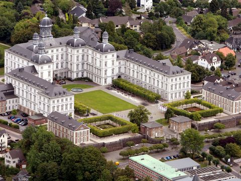 Luftaufnahme eines weißen Schlosses in U-Form gebaut. Im Innenhof befindet sich eine Rasenfläche. Das Schloss ist umgeben von Wohnbebauung. 