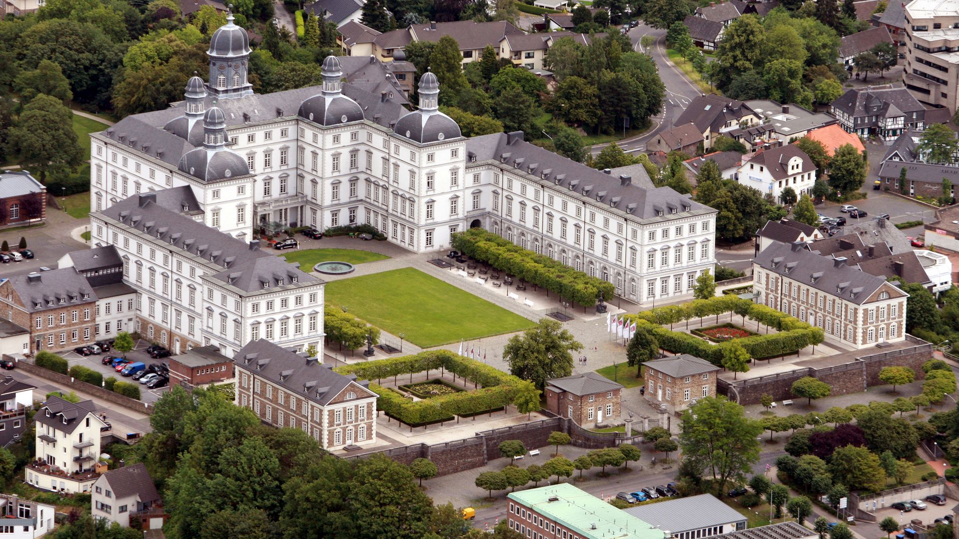 Das Foto zeigt eine Luftaufnahme von Bensberg mit dem Schloss Bensberg in der Mitte. Die weiße U-Form des Schlosses nimmt den Großteil des Bildes ein. Ringsherum sieht man Häuser, Parkplätze und Straßen.