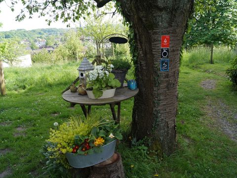 An einem Baum ist das Markierungszeichen des Obstweges angebracht, darunter steht ein kleiner runder Holztisch mit Blumen und einem Vogelhaus.