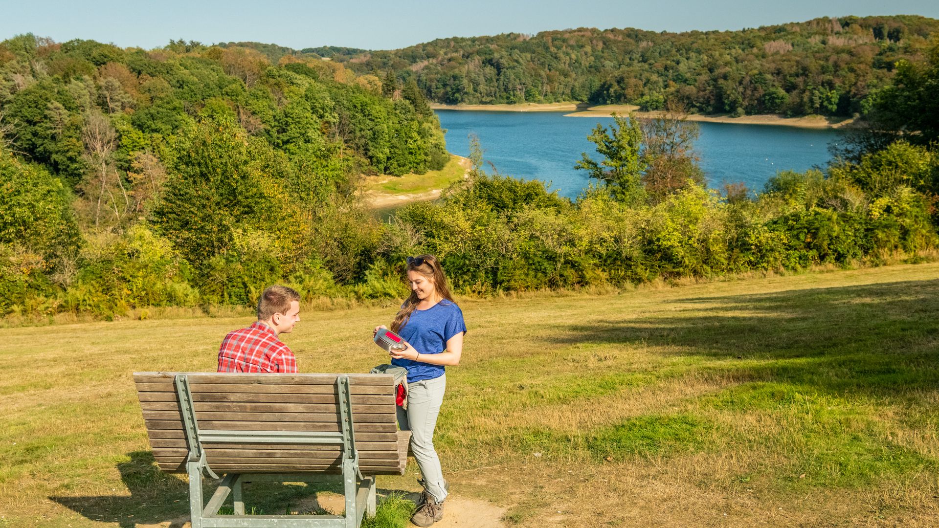 Ein junges Paar in Wanderausrüstung macht eine Pause mit Blick auf die Wahnbachtalsperre. Er sitzt auf einer Bank, sie steht danachen. 