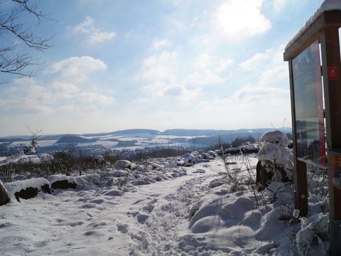 An einem Aussichtspunkt steht eine Informationstafel des Steinhauerpfades. Es liegt Schnee und die Sonne scheint. Der Blick reicht über den Steinbruch und über die Hügel des Bergischen Landes.