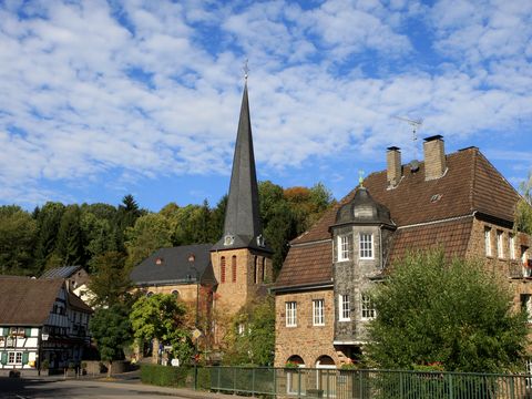 Eine gemauerte Kirche mit spitzem Kirchturm umgeben von Wohngebäuden und Bäumen. 