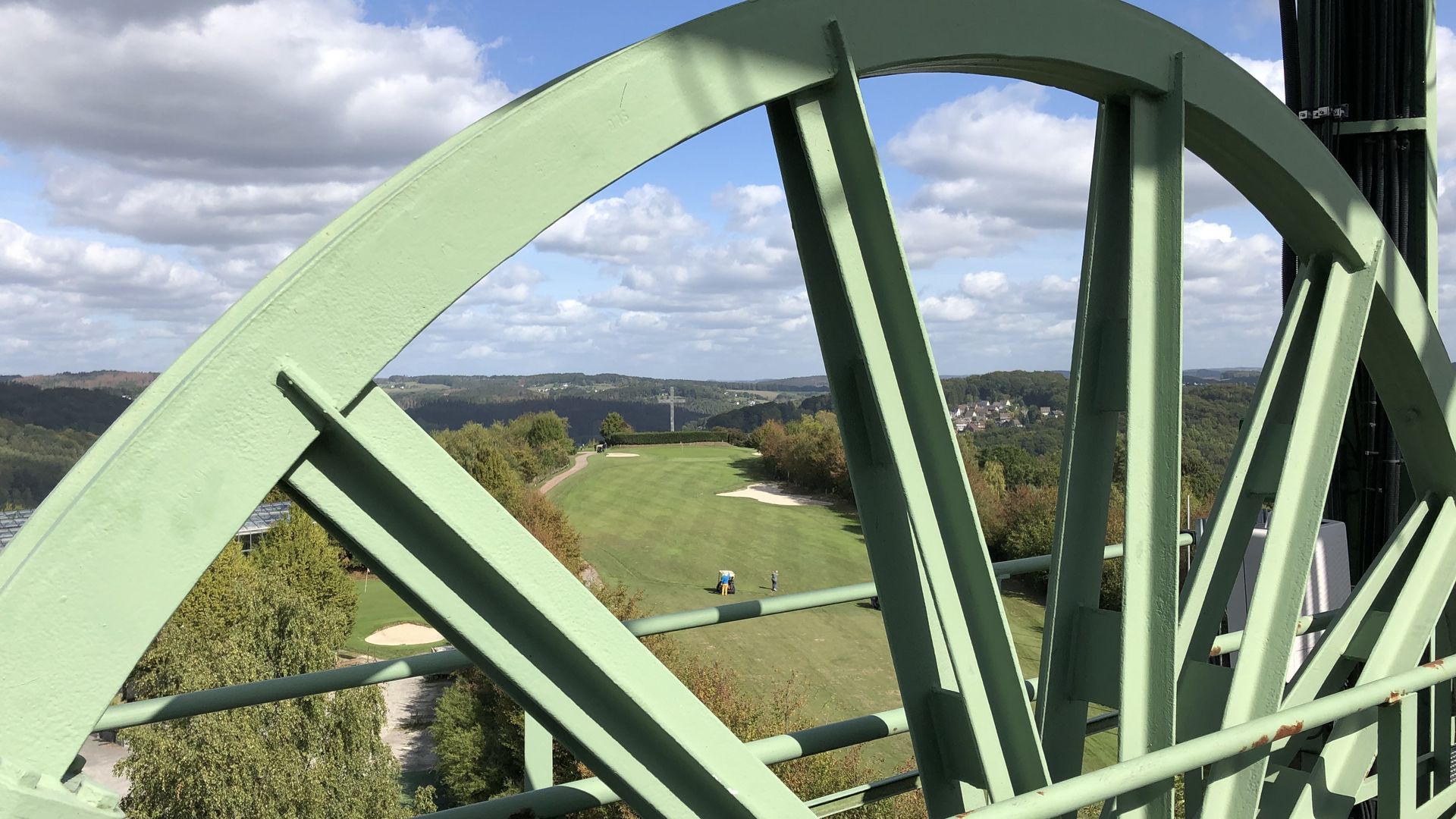 Im Vordergrund ist ein großes hellgrünes Rad vom Förderturm sichtbar. Vom Turm aus blickt man über einen Golfplatz