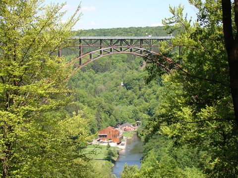 Blick auf das Tal unter der Müngestener Brücke mit einem Gebäude und einem Fluss. Links und rechts sind viele Bäume sichtbar, oben sieht man die Eisenbahnbrücke