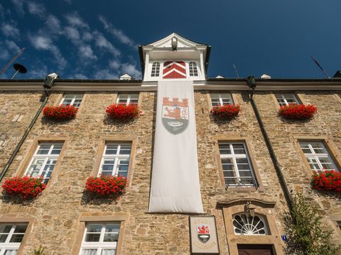 Blick vom Boden an einer Mauer von einem Schloss hoch in den blauen Himmel. Am Haus ist sind zwhlreiche Fenster mit roten Blumen davor zu sehen. An der Wand hängt eine lange weiße Fahne mit dem Stadtwappen und oben auf dem Dach ist eine Gaube zu sehen. 