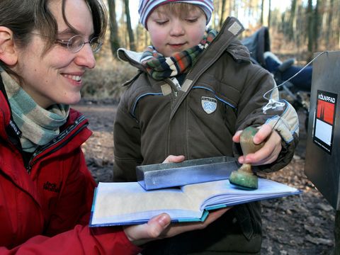 Eine Frau hält einen Block, damit ein etwa vierjähriges Kind einen Stempel auf den Block drücken kann. 