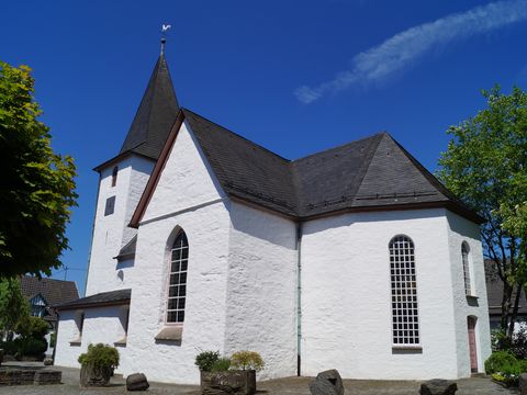 Das Foto zeigt die weiße Bonte Kerke in Lieberhausen von außen. Links und rechts daneben stehen grüne Bäume, der Himmel ist blau.