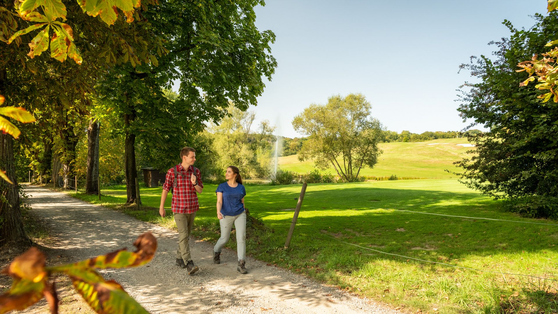 Ein junges Pärchen wandert auf einem flachen Wanderweg. Sie schauen sich an. Im Hintergrund ist ein Golfplatz und ein Springbrunnen zu sehen.