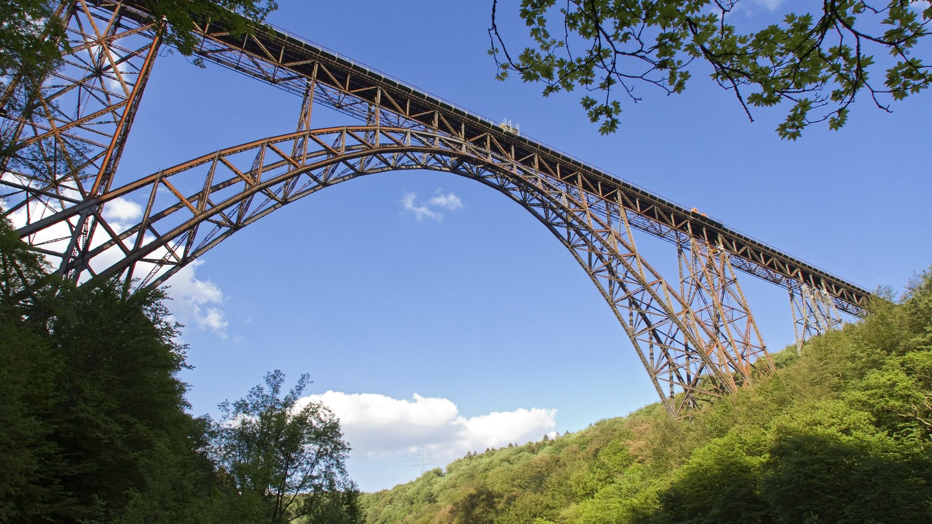 Blick vom Tal heraus auf die Müngstener Brücke vor blauem, wolkenlosen Himmel. Auf beiden Seiten der Brücke stehen Bäume. 