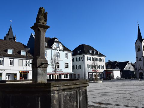 Blick auf einen gepflasterten Marktplatz mit einem Brunnen im Vordergrund und Häusern und einer Kirche im Hintergrund. 