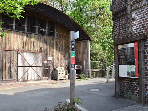 Zwischen einer alten Scheune und einem roten Backsteinhaus, von dem man rechts im Bild eine Wand sieht, steht eine Infotafel vom Tuchmacherweg und ein Wanderpfosten mit Richtungsweisern und Markierungszeichen.