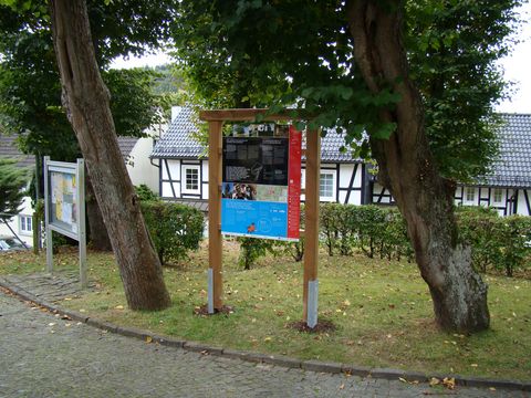 Am Wegesrand unter zwei Bäumen steht eine Infotafel vom Baumweg. Im Hintergrund kann man durch die Bäume hindurch Fachwerkhäuser erkennen.