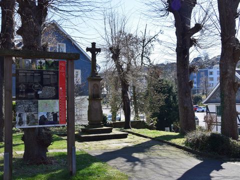 Eine Infotafel des Steinhauerpfades steht vor winterlich kahlen Bäumen und einem Denkmal, das ein Kreuz darstellt.