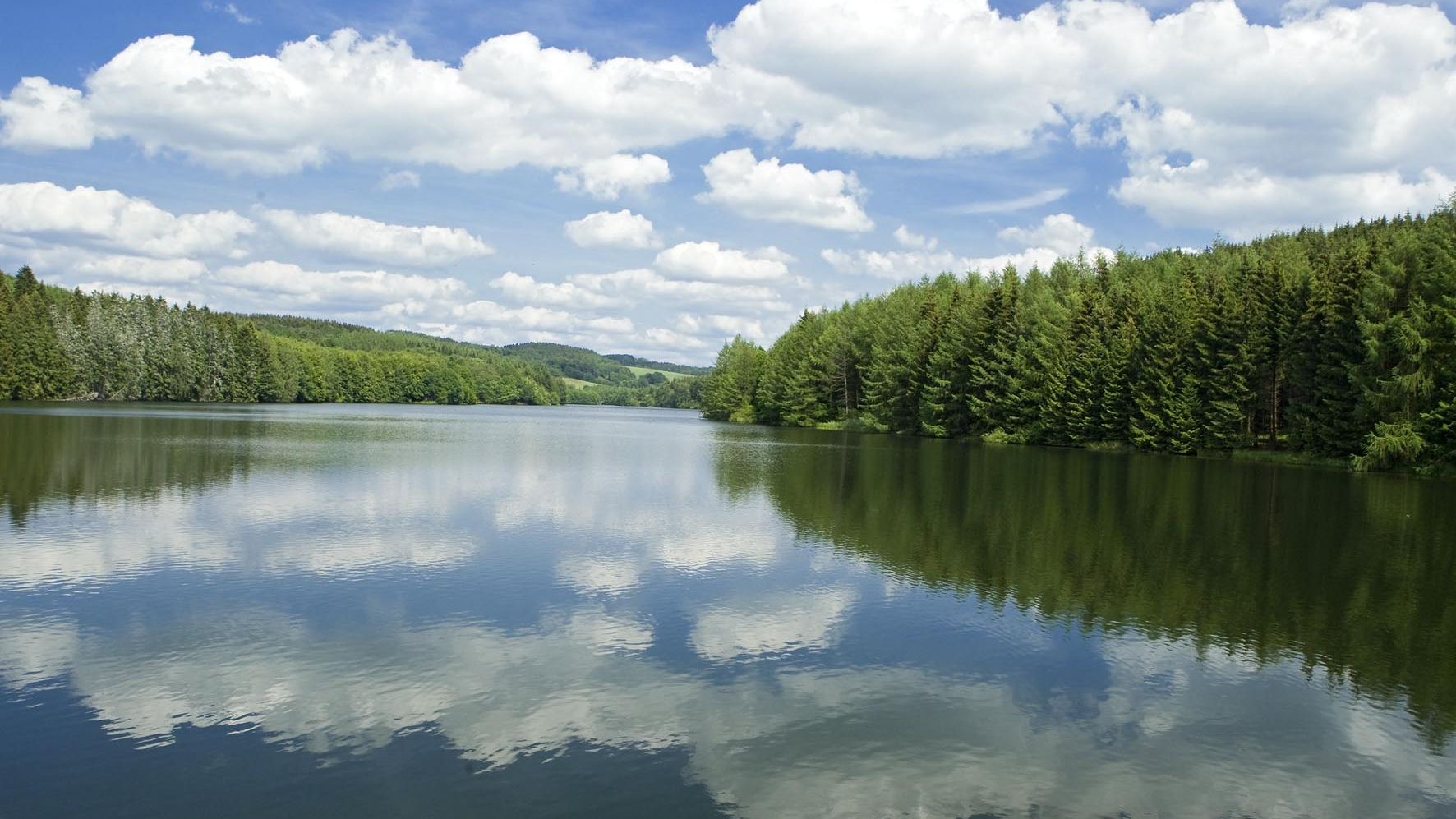 Wasserfläche einer Talsperre in der sich die weißen Wolken spiegeln. An den Ufern stehen Bäume