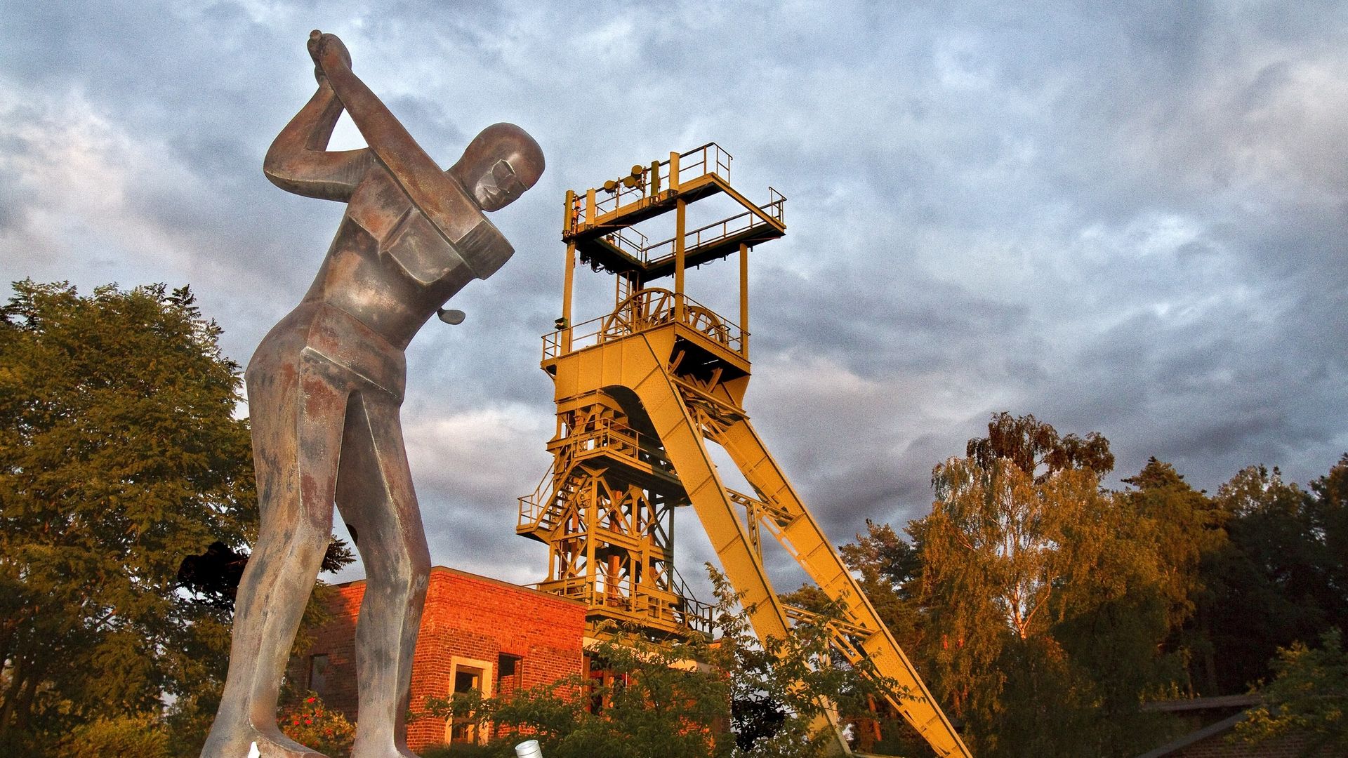 Im Vordergrund steht eine Statue eines Golfspielers, die den Golfschläger schwingt. Im Hintergrund steht der gelbe Förderturm am Golfplatz Lüderich.