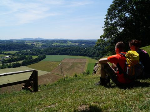 Ein junges Pärchen mit Wanderausrüszung sitzt auf einer Wiese auf einem Hügel und blickt auf die weite, offene Landschaft. 