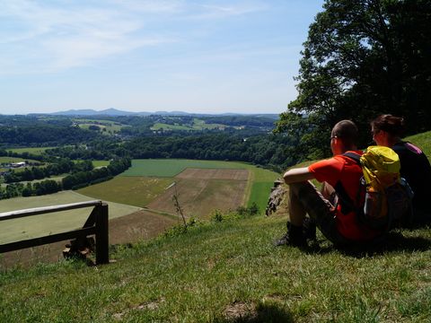 Ein junges Pärchen mit Wanderausrüszung sitzt auf einer Wiese auf einem Hügel und blickt auf die weite, offene Landschaft. 