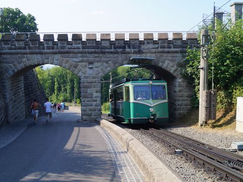 Ein grüner Zug fährt unter einer einer Brücke durch auf den Fotografen zu. Gleichzeit spazieren zwei Menschen auf dem Bahnsteig neben dem Zug durch einen zweiten Bogen der Brücke