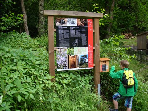 Inmitten grüner Sträucher steht eine Infotafel des Bergbauweges. An der daran angebrachten Audiostation kurbelt ein Kind in grüner Jacke und mit gelbem Rucksack an der Kurbel.