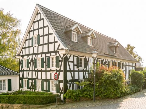 Fachwerkhaus mit schwarzen Balken, weißem Gefache und grünen Schlagläden mit einer grünen Hecke davor in einer Straße mit Kopfsteinpflaster