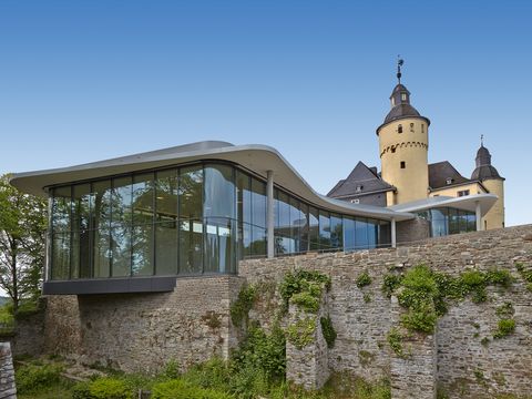 Blick auf eine etwas bewachsene Mauer hinter der zuerst ein moderner Anbau aus Glas und dann ein gelbes Schloss zu sehen ist. 